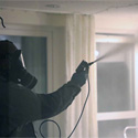 Ein Schädlingsbekämpfer steht in einem Raum, in voller Schutzausrüstung, bestehend aus Schutzmaske, Einwegschutzanzug und Handschuhen und führt eine Desinfektionsmaßnahme durch.